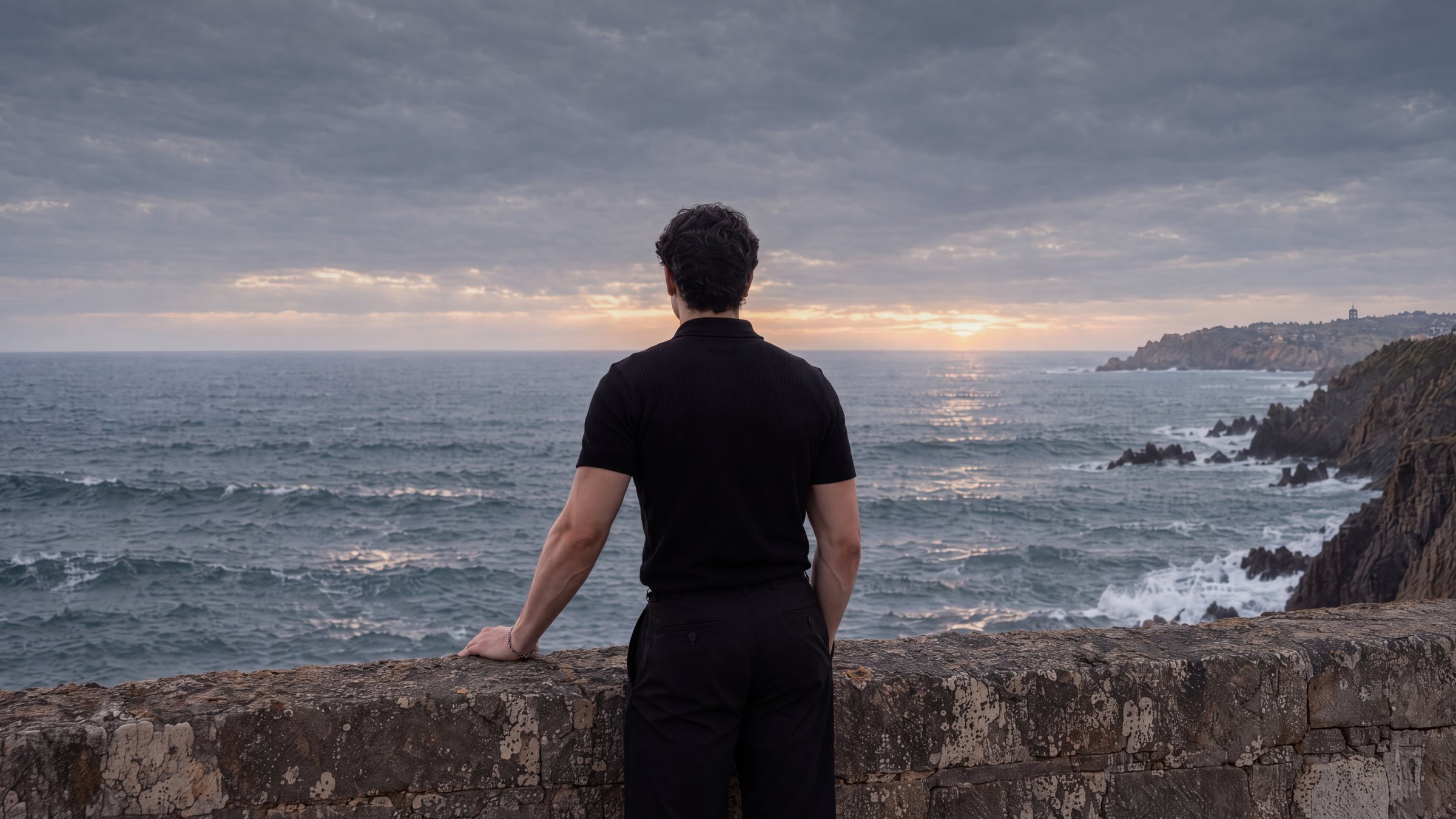 Arnav Jamwal, seen from behind, looking out over the sea from a stone ledge at dusk